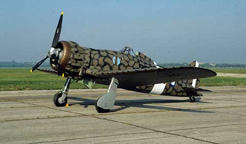 This is a vintage single-propeller military aircraft with camouflage paint, parked on a concrete ground against a clear sky. It has distinct roundel insignia.