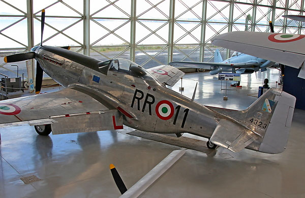 This image shows a vintage metallic fighter aircraft with identification "RR011" inside a hangar with large geometric windows and other aircraft in the background.