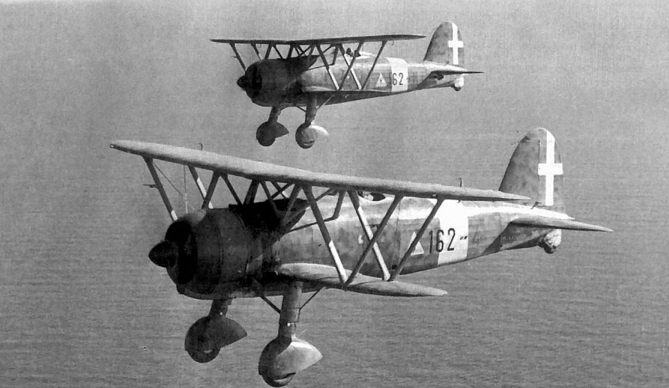 This is a black and white photo of two vintage biplanes in flight, with identifiable markings and landing gear visible, against a hazy background.