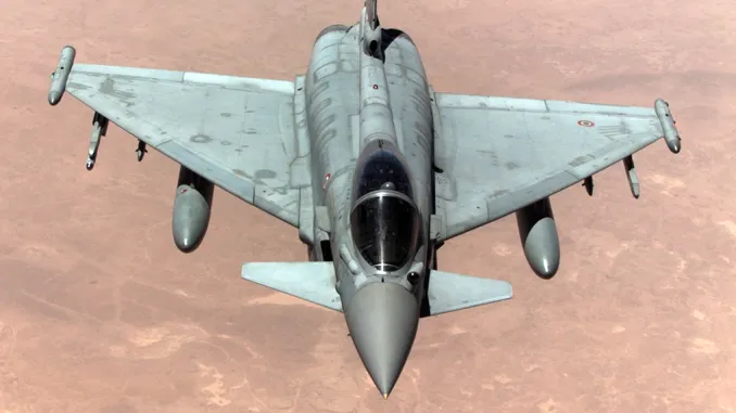 The image shows a military jet in-flight, seen from above, against a sandy, desert-like background. It has two wing-mounted drop tanks and missiles.