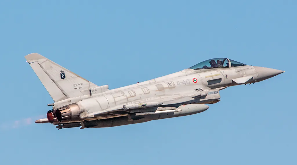 A military fighter jet in flight against a clear blue sky, with visible armaments and the afterburner glowing, indicating high speed. A pilot is discernible in the cockpit.